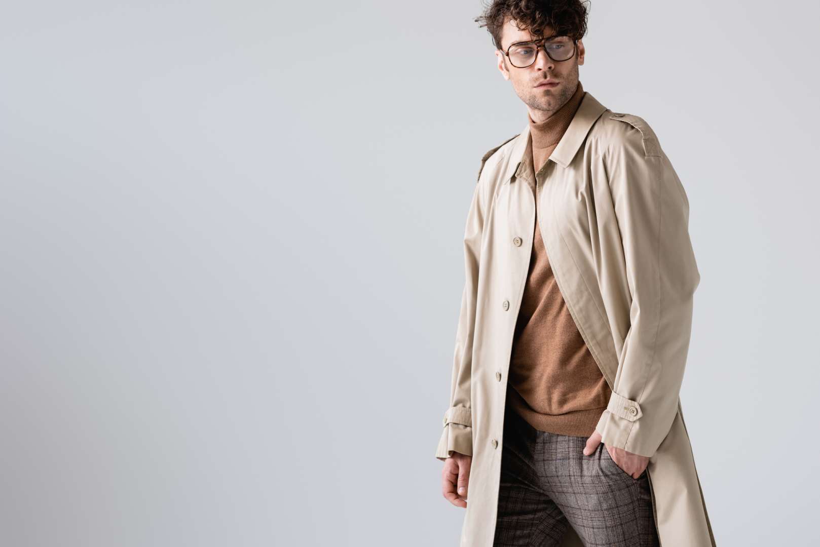 Young man with tousled hair and glasses in a trench coat, sweater, and patterned pants