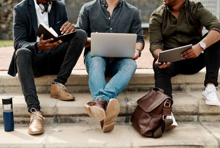 A group of men, wearing a variety of shoe styles, sitting on stairs around a laptop