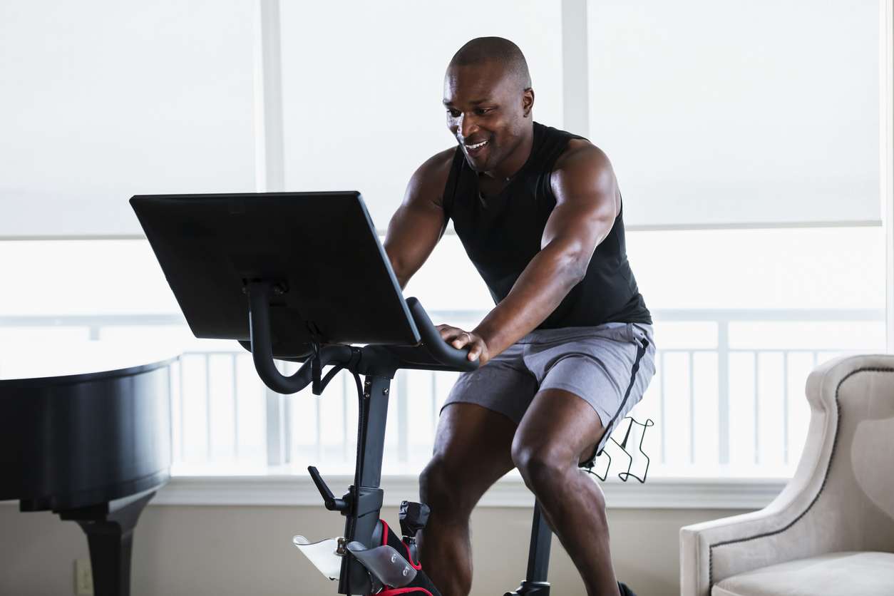 An adult African-American man in his 30s exercising at home in his living room, on a Peloton bike.