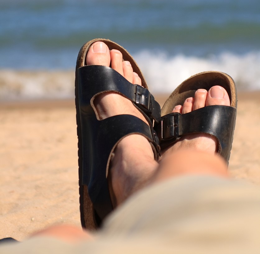 A man wearing a pair of classic Birkenstock-style sandals lounges on the beach 